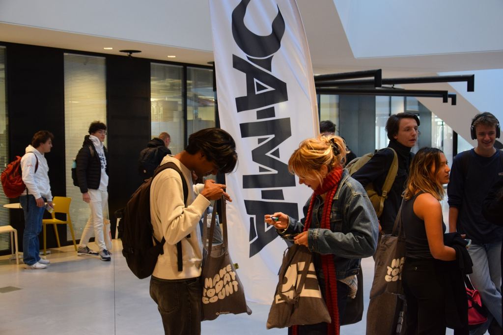 Le stand de l’AMAC dans un hall de l’EPFL : son drapeau, et des étudiant·e·s qui découvrent leur sac de bienvenue